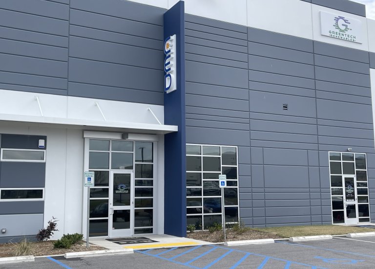 Side angled front view of Greentech Renewables building. The building is grey with blue and white accents. There are large "CED" letters on the ledge and a full color logo with "Greentech Renewables" on the front and doors.